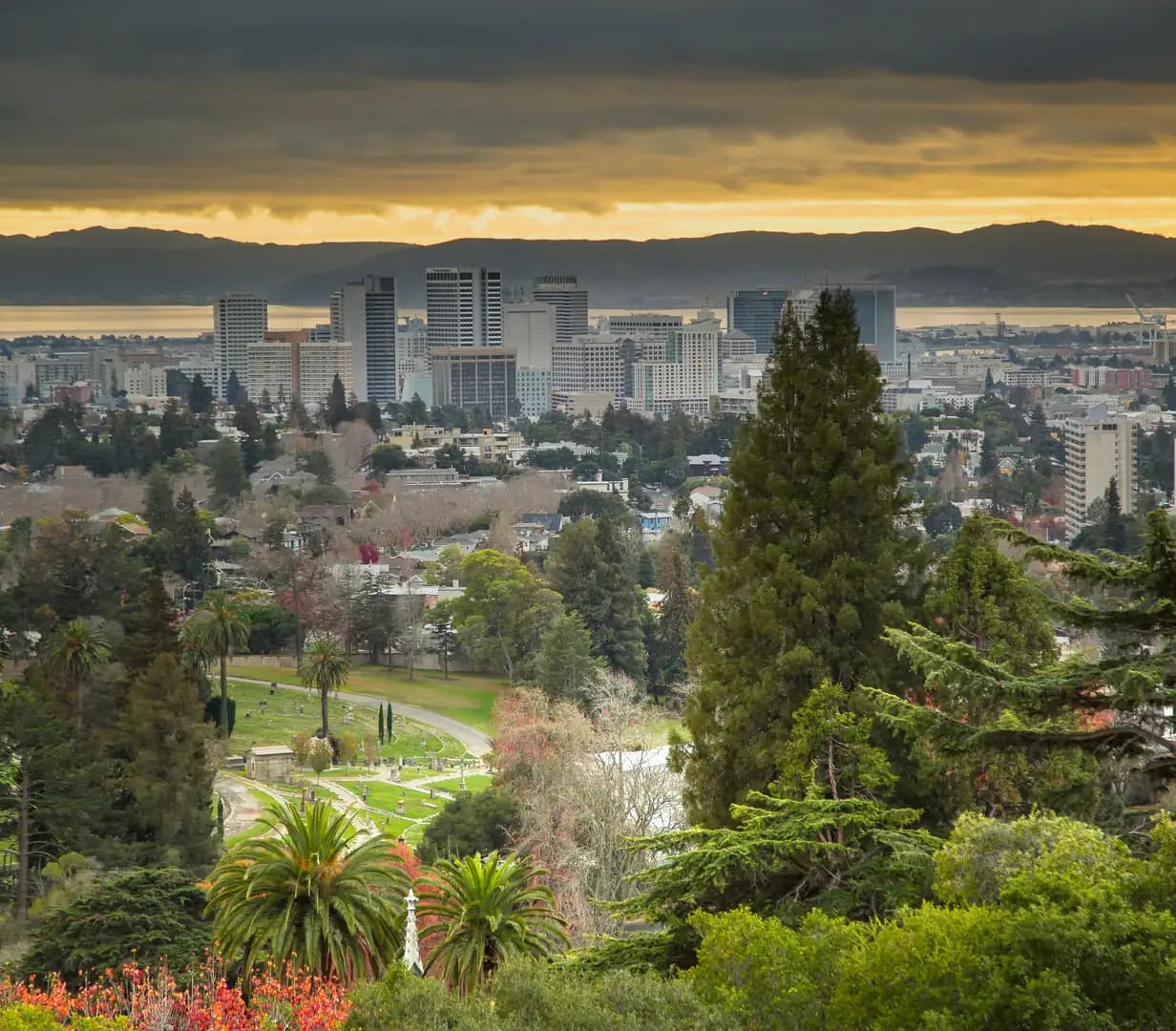 Downtown Oakland, California skyline and San Francisco Bay seen over green hillside trees under a golden evening sky