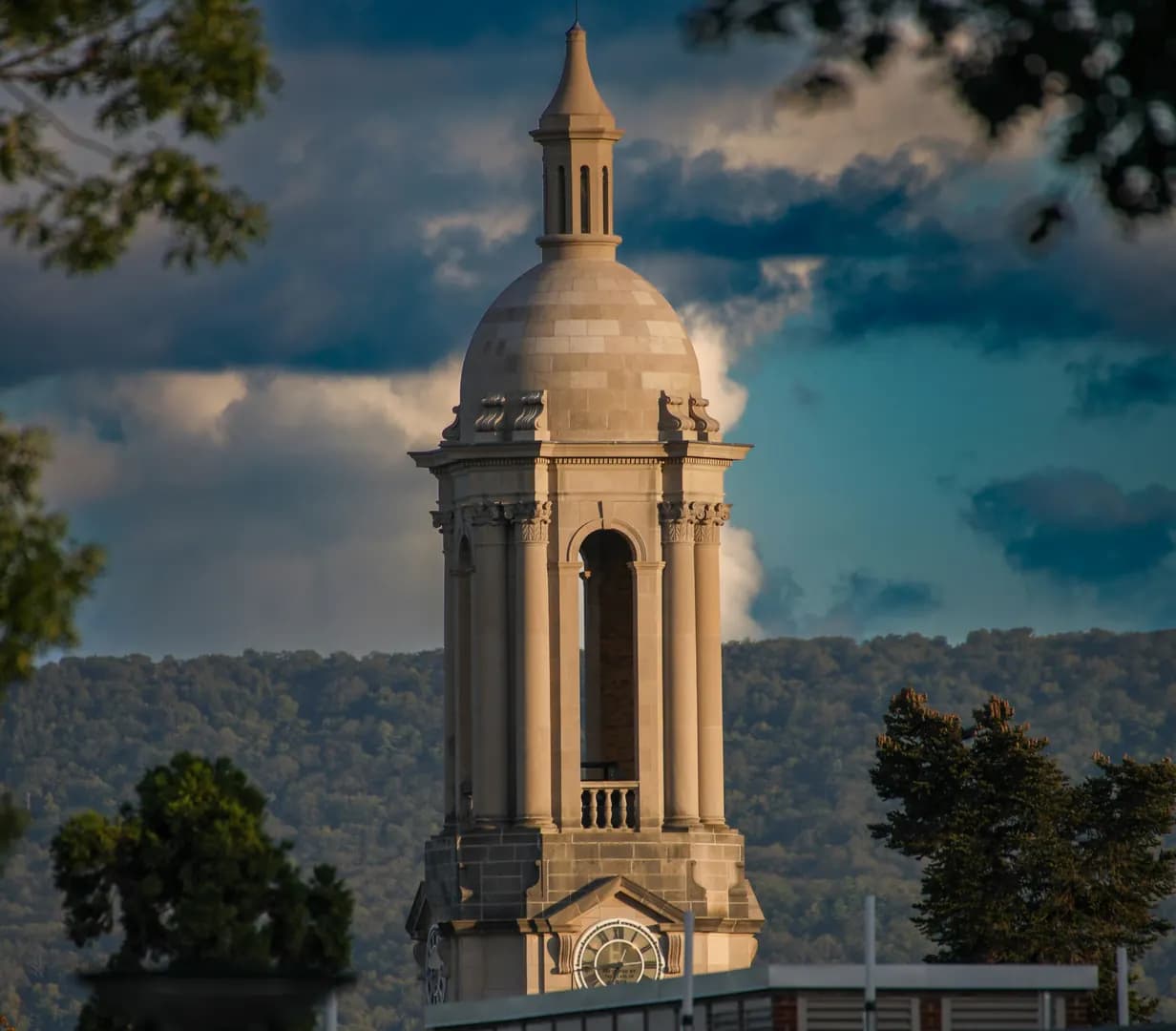 Old Main bell tower at Penn State in State College, Pennsylvania, with a wooded ridge and storm clouds behind