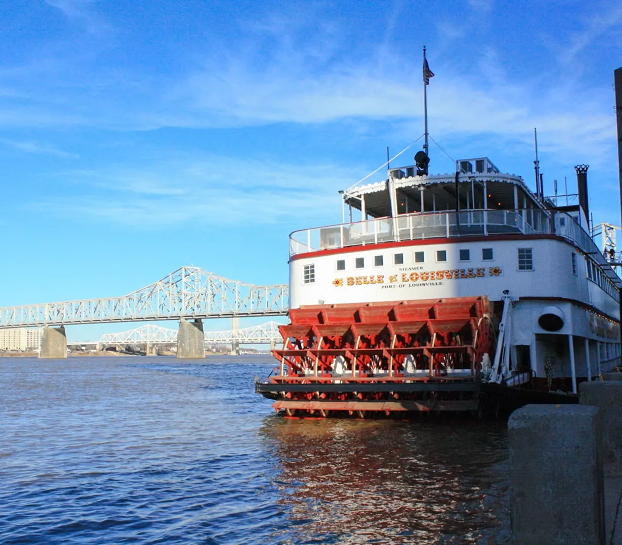 Belle of Louisville paddlewheel steamboat on the Ohio River below a Louisville, Kentucky bridge, a stop for charter bus groups